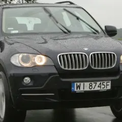 Ciemny BMW E70 na mokrej drodze, w tle zielone pola i drzewa.