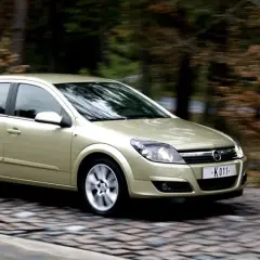 Złoty Opel Astra H 1.6 benzyna w ruchu na brukowanej drodze, otoczony jesiennym lasem.
