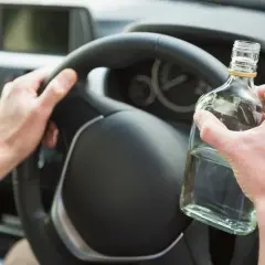 Ręce kierowcy na kierownicy, jedna trzyma butelkę alkoholu. Nie warto sprawdzać, ile promili.