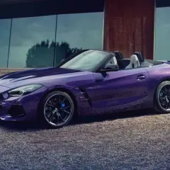 Fioletowy roadster BMW Z4 z otwartym dachem, zaparkowany na żwirowej nawierzchni przed nowoczesnym budynkiem.