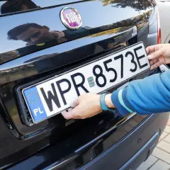 Ręce montują tablicę rejestracyjną WPR 8573E na czarnym samochodzie. To duplikat tablicy rejestracyjnej.