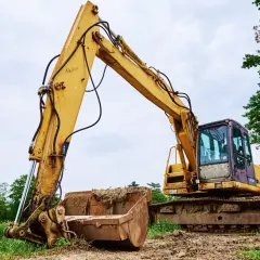Żółta koparka z pełnym łyżką ziemi, gotowa do pracy. Widać jej gąsienice i mechanizm podnoszenia ramienia, a także zwolnica.