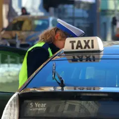 Kontroler drogowy w kamizelce odblaskowej pochyla się nad taksówką, sprawdzając dokumenty. Może to oznaczać mandat za brak pasów.