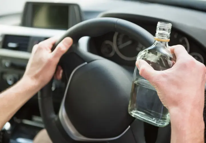 Ręce kierowcy na kierownicy, jedna trzyma butelkę alkoholu. Nie warto sprawdzać, ile promili.