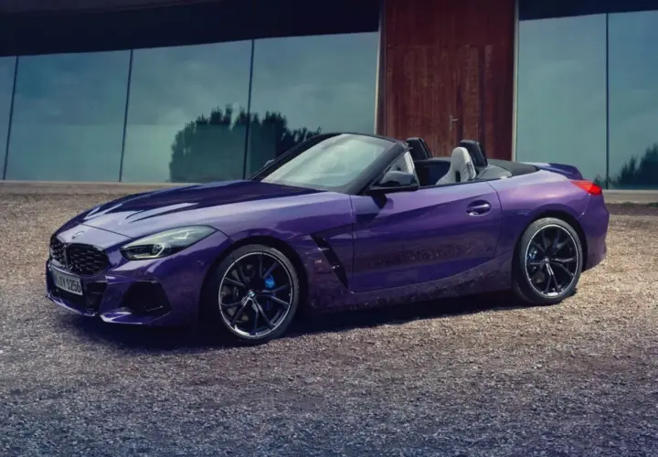 Fioletowy roadster BMW Z4 z otwartym dachem, zaparkowany na żwirowej nawierzchni przed nowoczesnym budynkiem.
