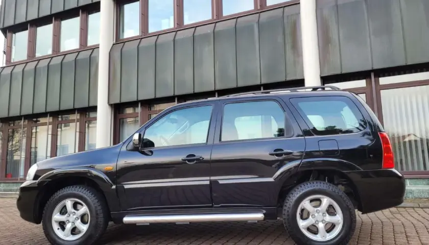Czarny Mazda Tribute stoi na brukowanym podjeździe przed nowoczesnym budynkiem.