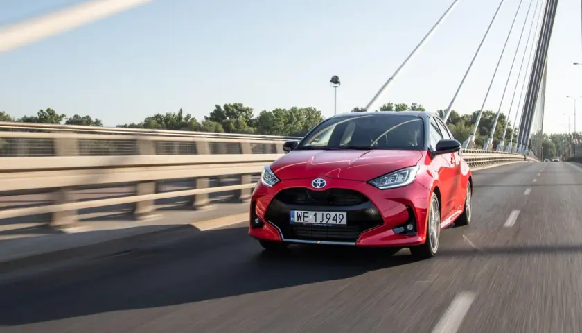 Czerwony Toyota Yaris Hybrid pędzi po moście w słoneczny dzień.