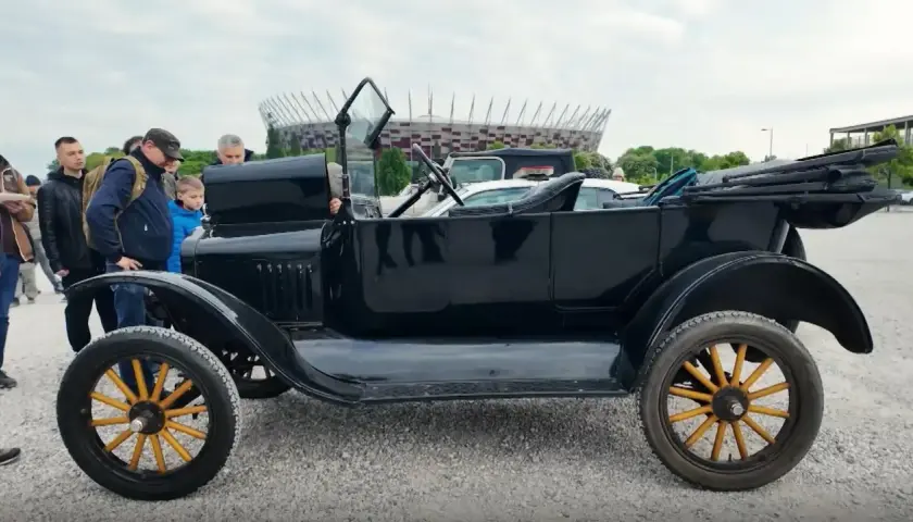 Czarny, zabytkowy Ford T z drewnianymi szprychami kół, obok którego stoją ludzie. W tle widać stadion.