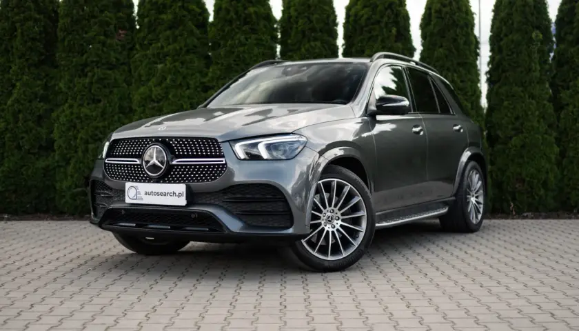 Elegancki, szary Mercedes GLE 400d SUV stoi na wybrukowanym podjeździe, otoczony gęstymi, zielonymi żywopłotami.