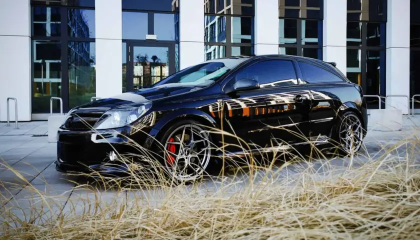 Czarny Opel Astra GTC z chromowanymi felgami i czerwonymi zaciskami hamulcowymi, zaparkowany przed nowoczesnym budynkiem, otoczony suchą trawą.