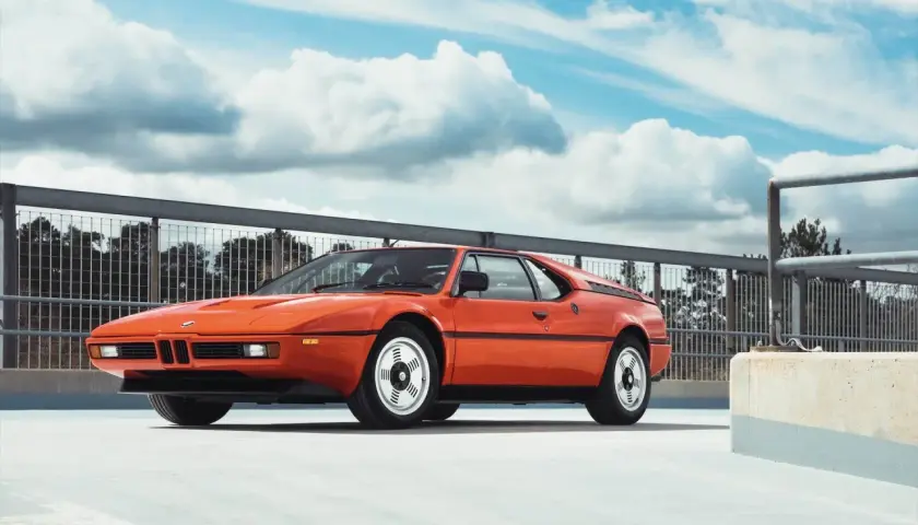 Pomarańczowe BMW E26, ikona lat 80., prezentuje się na tle błękitnego nieba z chmurami.