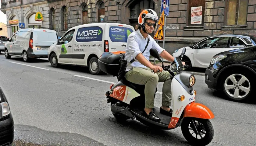 Mężczyzna w kasku jedzie skuterem elektrycznym, posiadając prawo jazdy AM. W tle białe vany dostawcze.