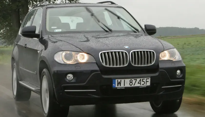Ciemny BMW E70 na mokrej drodze, w tle zielone pola i drzewa.