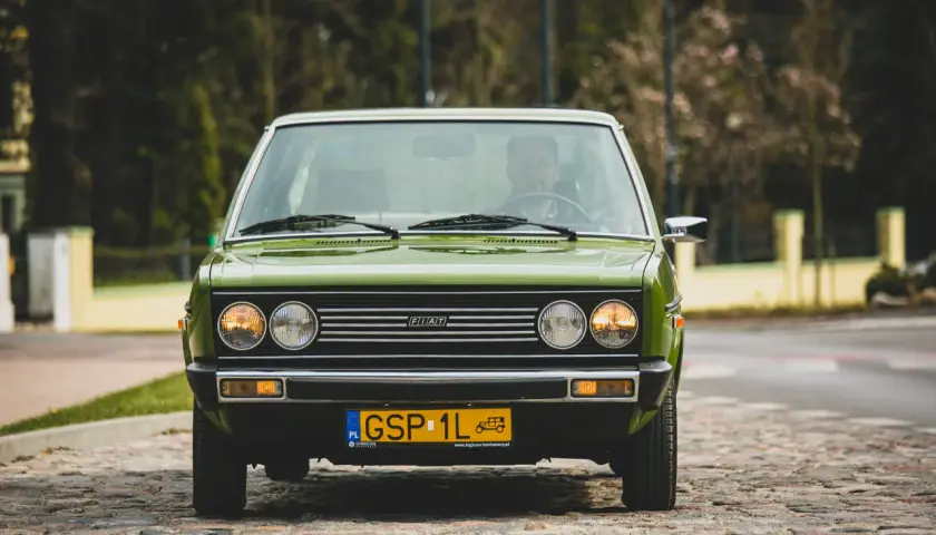 Zielony Fiat 131 Mirafiori z lat 70. na brukowanej ulicy, z widocznym kierowcą za kierownicą.