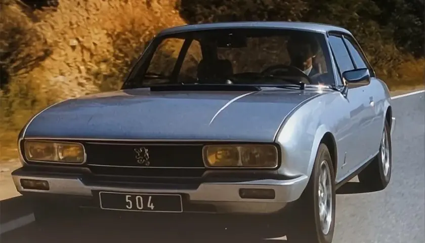 Srebrny Peugeot 504 z lat 70. jedzie po krętej drodze wśród wzgórz.