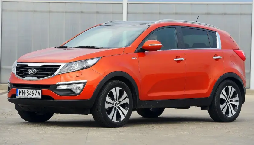Pomarańczowy Kia Sportage 2015 z napędem AWD, zaparkowany na betonowym placu przed budynkiem z przeszkloną fasadą.