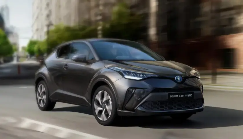Ciemnoszary Toyota C-HR Hybrid w ruchu miejskim, z rozmytym tłem budynków i drzew.