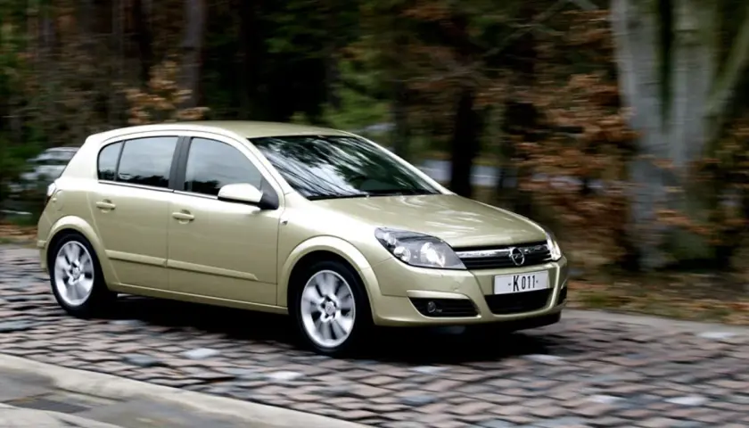 Złoty Opel Astra H 1.6 benzyna w ruchu na brukowanej drodze, otoczony jesiennym lasem.