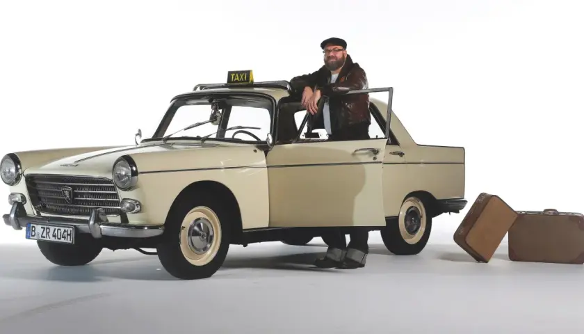 Mężczyzna w skórzanej kurtce opiera się o zabytkowy Peugeot 404 z tabliczką TAXI. Obok stoją walizki.
