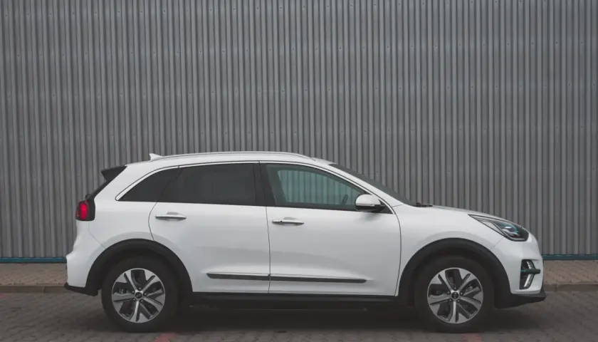Biały Kia Niro EV stoi na tle szarej, falistej ściany. Elegancki design i nowoczesne linie podkreślają jego ekologiczny charakter.