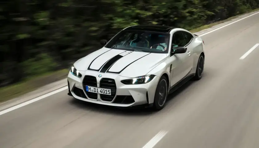 Białe BMW F82 z czarnymi pasami na masce pędzi po krętej drodze wśród zieleni.