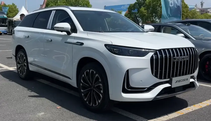 Biały SUV Jaecoo 9 z chromowanym grillem i czarnymi felgami stoi na parkingu.