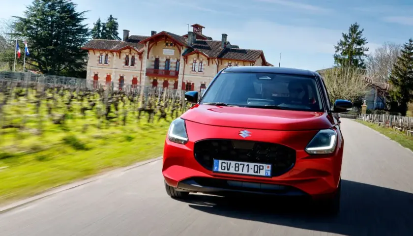 Czerwony Suzuki Swift pędzi drogą obok winnicy i zabytkowego domu.