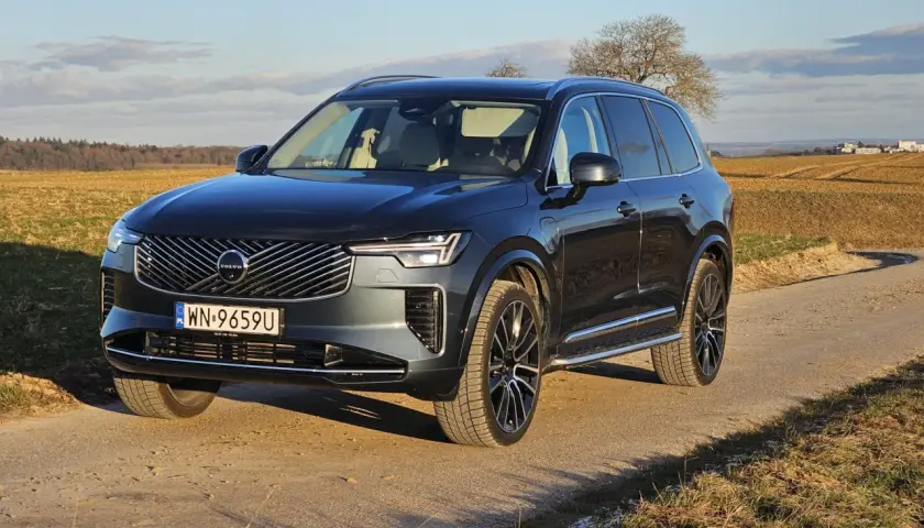 Ciemnoszary Volvo XC90 T8 na drodze polnej, z polami i drzewami w tle.