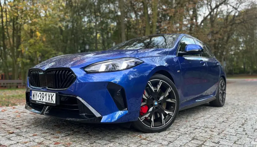 Błękitne BMW 235i z czarnymi felgami i czerwonymi zaciskami hamulcowymi zaparkowane na bruku wśród jesiennych drzew.