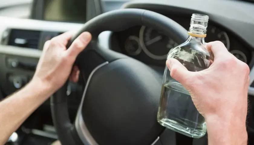 Ręce kierowcy na kierownicy, jedna trzyma butelkę alkoholu. Nie warto sprawdzać, ile promili.