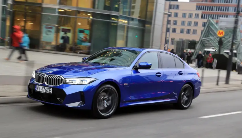 Błękitne BMW 330e mija nowoczesne budynki i wejście do metra.