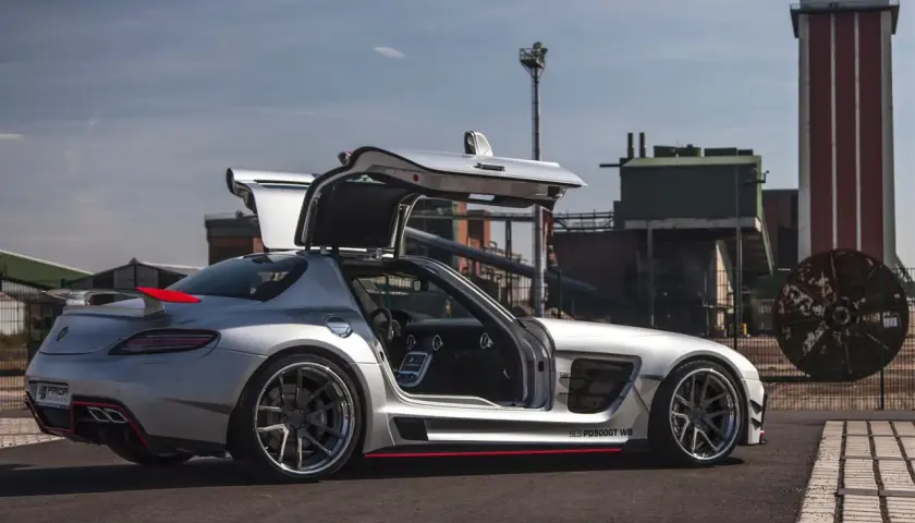Srebrny SLS z otwartymi drzwiami typu gullwing, z czerwonym akcentem na spojlerze, zaparkowany przed industrialnym budynkiem.