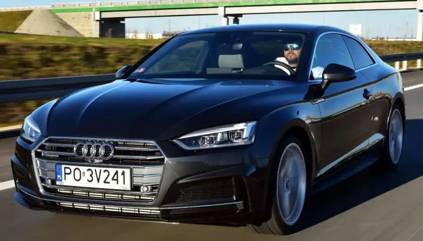 Czarny Audi A5 2025 z kierowcą w okularach przeciwsłonecznych jedzie po autostradzie.