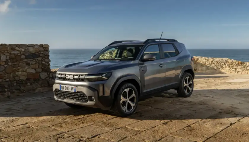 Nowy Dacia SUV na tle kamiennego muru i morza.