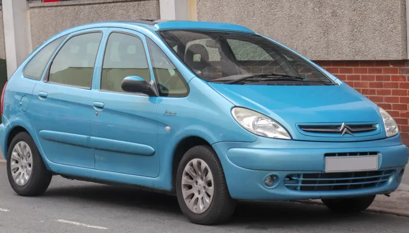 Błękitny Citroen Xsara Picasso z napisem "Picasso" na błotniku, zaparkowany przy ceglanej ścianie.
