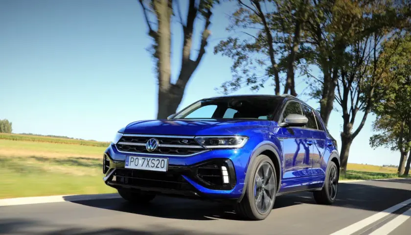 Niebieski Volkswagen T-Roc R pędzi po wiejskiej drodze, otoczony drzewami i polami.