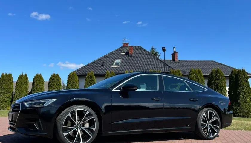 Elegancki czarny Audi A7 C8 lśni w słońcu na tle domu i zielonych drzew.