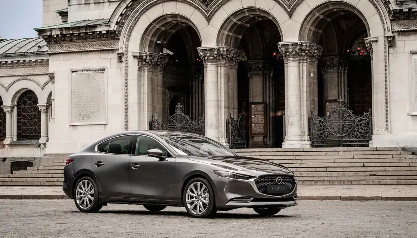 Elegancka, szara Mazda 3 2025 przed monumentalną, kamienną budowlą z arkadami i ozdobnymi bramami.