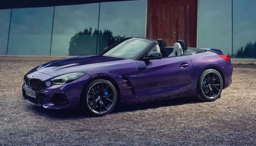 Fioletowy roadster BMW Z4 z otwartym dachem, zaparkowany na żwirowej nawierzchni przed nowoczesnym budynkiem.