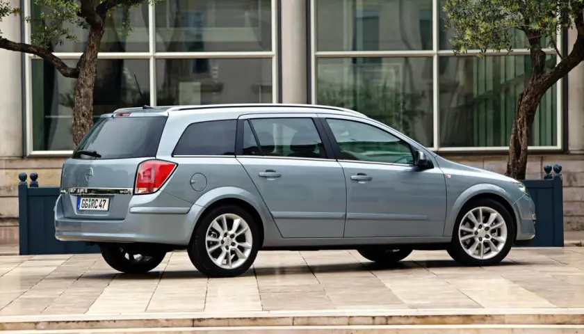 Srebrny Opel Astra 2005 kombi zaparkowany przed nowoczesnym budynkiem z drzewami.