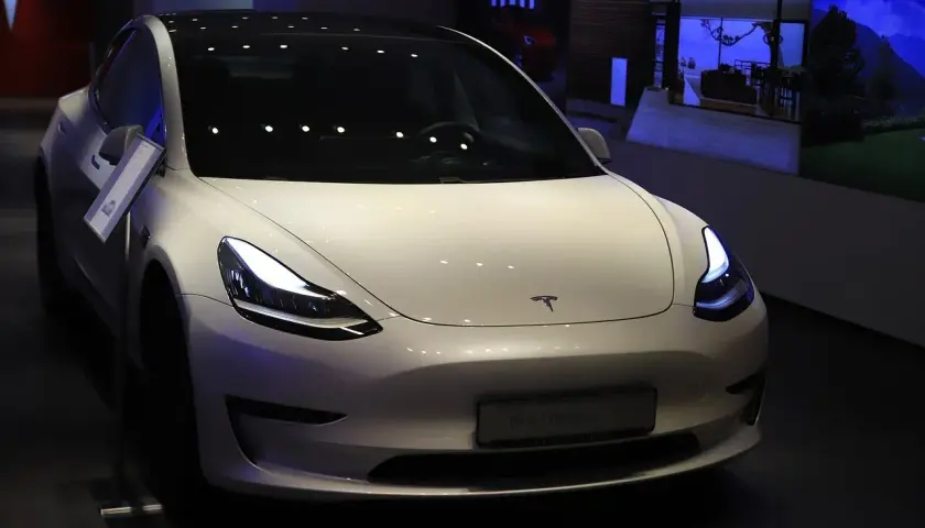 Biały samochód Tesla Model 3 z włączonymi światłami, prezentowany w salonie.