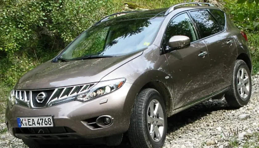 Brązowy Nissan Murano na kamienistej drodze, otoczony zielenią.