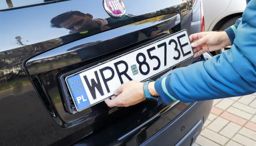 Ręce montują tablicę rejestracyjną WPR 8573E na czarnym samochodzie. To duplikat tablicy rejestracyjnej.