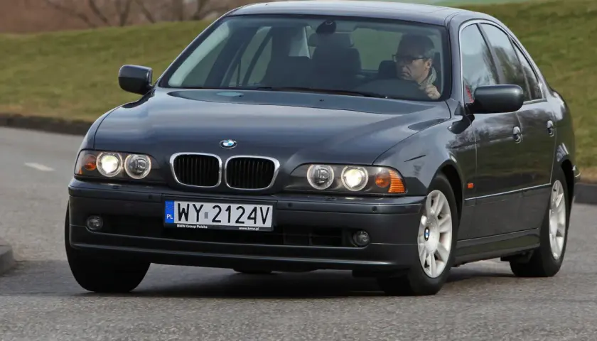 Ciemnoszary sedan BMW E39 z kierowcą w okularach jedzie po drodze.