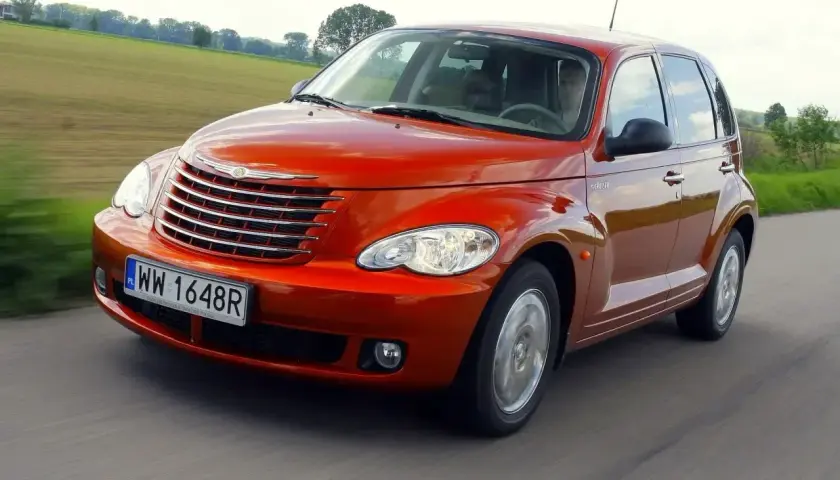 Pomarańczowy Chrysler PT Cruiser jedzie po drodze, za nim zielone pola i drzewa.