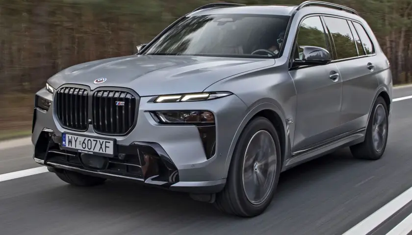 Szary BMW X7 2025 z czarnym grillem i sportowymi akcentami pędzi po drodze wśród drzew.
