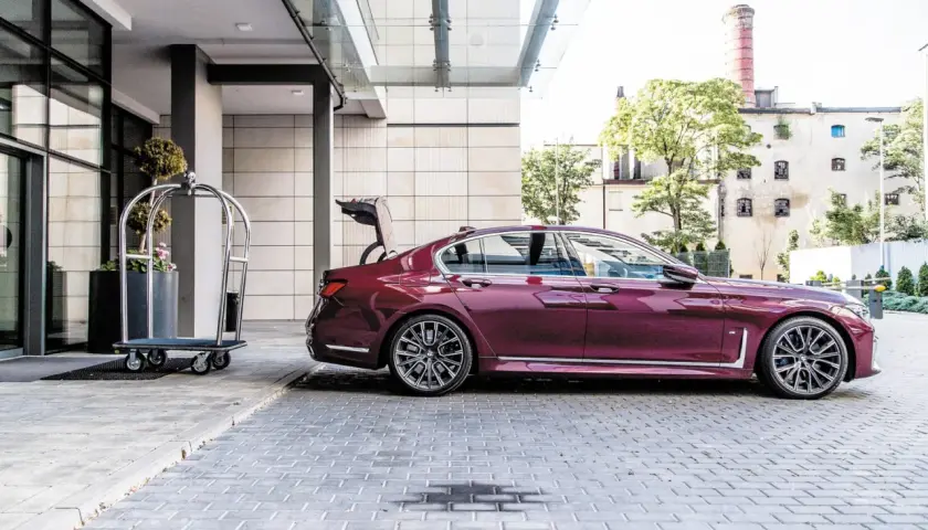 Eleganckie bmw g11 w kolorze śliwkowym z otwartym bagażnikiem stoi przed hotelem obok wózka na bagaż.
