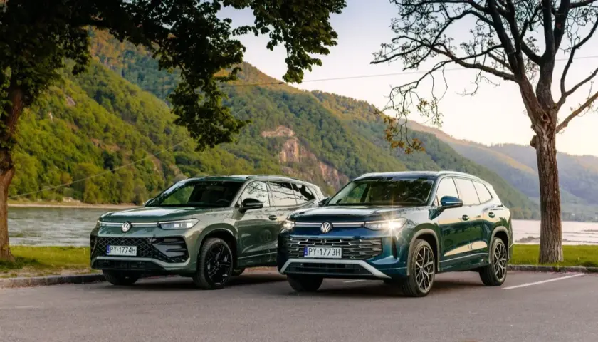 Dwa nowe VW Tayron, zielony i granatowy, zaparkowane nad rzeką, z górami w tle.