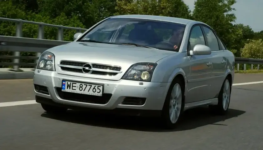 Srebrny Opel Vectra C GTS pędzi po drodze, otoczony zielenią.