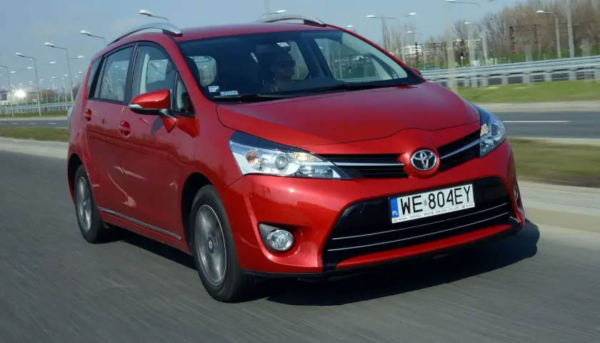 Czerwona Toyota Corolla Verso jedzie po drodze. Kierowca w okularach przeciwsłonecznych prowadzi auto.